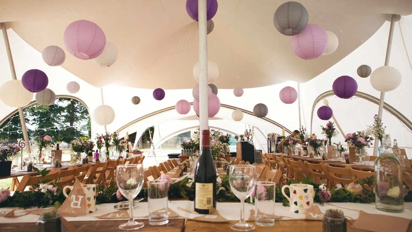 Rustic tables and folding wooden chairs dressed under paper lanterns in a marquee