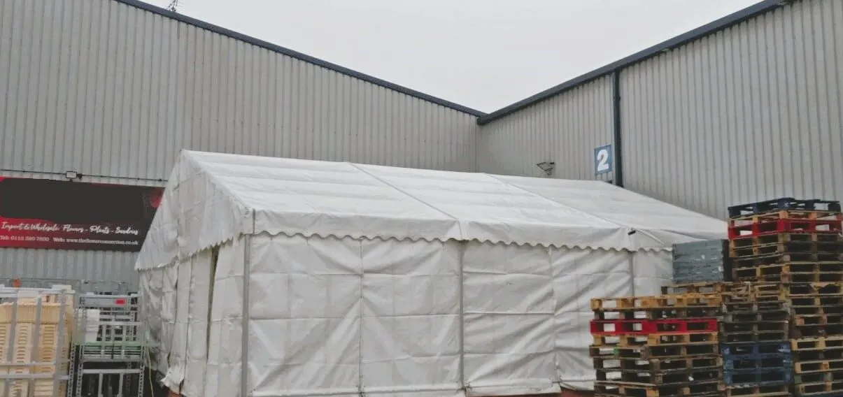 A walled frame marquee on hard ground beside an industrial unit, stacked pallets alongside